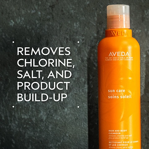 Image 2 - An orange Aveda Sun Care hair and body cleanser bottle with a translucent white cap is displayed on a dark grey textured background, with text on the left stating REMOVES CHLORINE, SALT, AND PRODUCT BUILD-UP, and bottle labels that read AVEDA THE ART AND SCIENCE OF PURE FLOWER AND PLANT ESSENCES sun care soins soleil HAIR AND BODY CLEANSER removes chlorine and salt plus product build-up NETTOYANT POUR LE CORPS ET LES CHEVEUX élimine chlore, sel et autres accumulation de produits. Image 3 - A dark green graphic with a white circle in the center containing three brown babassu nuts, with white text at the top reading BABASSU-DERIVED CLEANSING SYSTEM and at the bottom reading HELPS TO REMOVE CHLORINE AND SALT. Image 4 - Two orange Aveda sun care product bottles stand on a dark surface, with the larger bottle on the left and a smaller spray bottle on the right, accompanied by descriptive text on how to use them: HOW TO USE, 01 DEFEND Sun Care Protective Hair Veil, 02 CLEANSE Sun Care Hair and Body Cleanser, AVEDA THE ART AND SCIENCE OF PURE FLOWER AND PLANT ESSENCES, sun care soins soleil, HAIR AND BODY CLEANSER removes chlorine and salt plus product build-up, NETTOYANT POUR LE CORPS ET LES CHEVEUX élimine chlore, sel et autres accumulations de produits, 8.5 fl oz/oz liq/250 ml e, PROTECTIVE HAIR VEIL UV filters help protect hair, VOILE PROTECTEUR POUR LES CHEVEUX filtres anti-UV pour la protection des cheveux, 3.4 fl oz/oz liq/100 ml e. Image 5 - A puddle of light yellow, foamy liquid is shown on a white background. Image 6 - A background of dark green plant leaves with a red stem on the left shows three white certification logos: 100% VEGAN circling a V, Certified B Corporation with a B inside a circle, and Cruelty Free INTERNATIONAL featuring a leaping rabbit and stars with a TM.