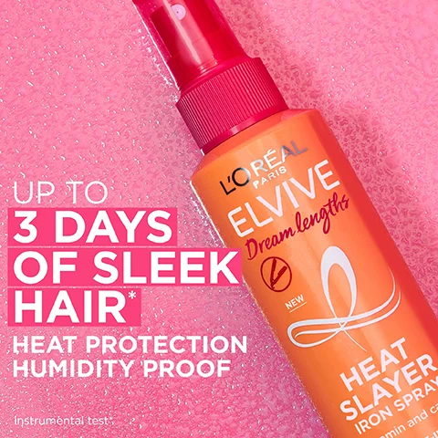 A close-up of an orange and pink LOreal Elvive Dream Lengths Heat Slayer Iron Spray bottle on a pink sparkly background with text that says UP TO 3 DAYS OF SLEEK HAIR* HEAT PROTECTION HUMIDITY PROOF instrumental test LOREAL PARIS ELVIVE Dream lengths NEW HEAT SLAYER IRON SPRAY vitamin and cast.